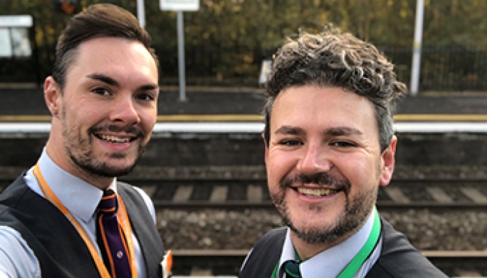 Sam and Dan smiling at the camera on a train platform.