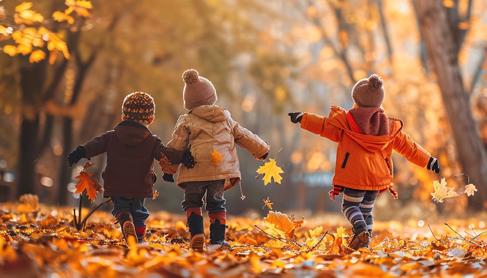 Three kids frolicking in a wooded area linking arms and tossing autumn foliage into the air