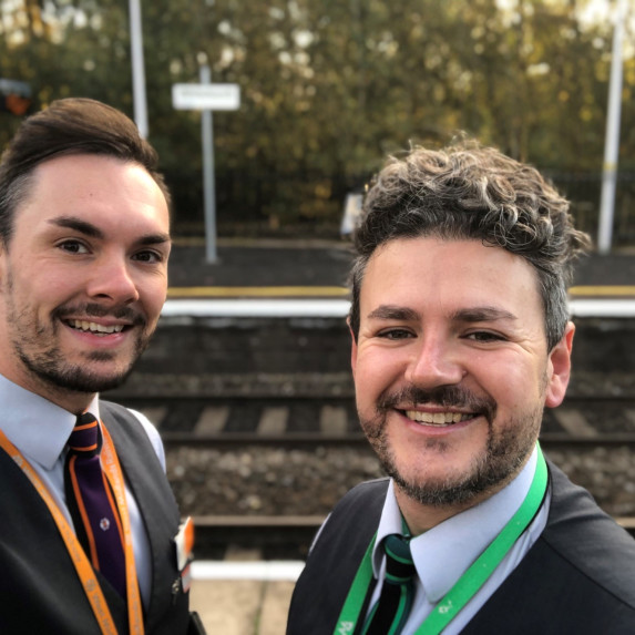 Sam and Dan smiling at the camera standing on a platform.